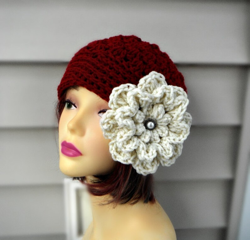 May include: A burgundy crochet hat with a large, cream-colored flower attached to the side. The flower has a pearl-like center. The hat is displayed on a mannequin head.