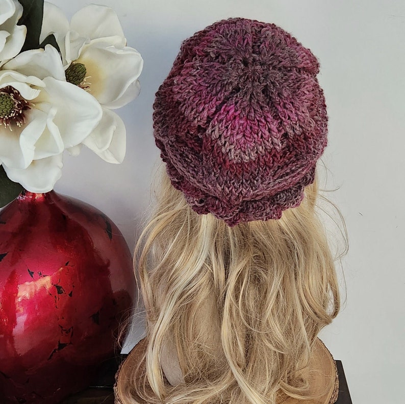 May include: A pink and purple knitted beanie with a textured pattern. The beanie is on a mannequin with long blonde hair.