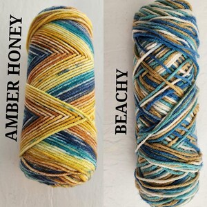 May include: Two skeins of yarn. The left skein is labeled AMBER HONEY and features a chevron pattern of yellow, gold, teal, and dark blue. The right skein is labeled BEACHY and features a variegated mix of white, tan, teal, and dark blue.