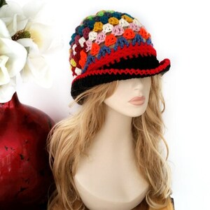 May include: A colorful crochet hat with a black brim and a red band. The hat features a patchwork design with squares of various colors, including blue, orange, yellow, and pink. The hat is displayed on a mannequin head with blonde hair.