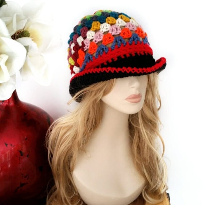 May include: A colorful crochet hat with a black brim and a red band. The hat features a patchwork design with squares of various colors, including blue, orange, yellow, and pink. The hat is displayed on a mannequin head with blonde hair.