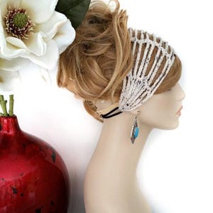 May include: A side profile of a mannequin head wearing a white crocheted headband. The mannequin also has a silver earring with a turquoise stone. A red vase with a white flower is in the background.