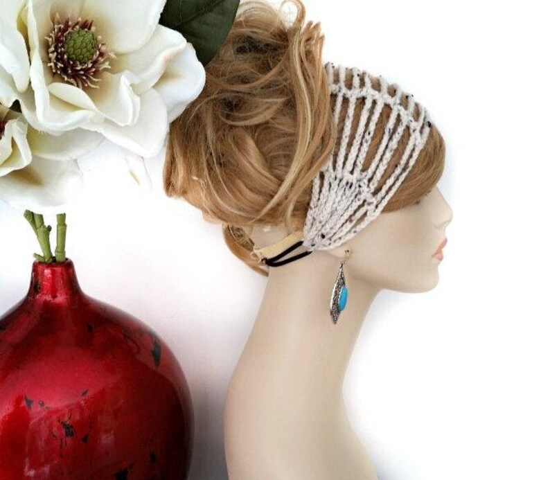 May include: A side profile of a mannequin head wearing a white crocheted headband. The mannequin also has a silver earring with a turquoise stone. A red vase with a white flower is in the background.