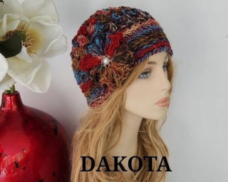 May include: A crocheted hat with a colorful floral design and a jeweled embellishment. The hat is made with a variety of shades of brown, red, blue, and green yarn. The hat is worn by a mannequin with long blonde hair. The word "DAKOTA" is printed in black letters on the bottom of the image.