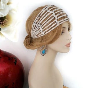 May include: A cream-colored, crocheted headband with a lattice design is worn on a mannequin with blonde hair. The mannequin also wears silver earrings with a turquoise stone. A red vase with white flowers is in the background.