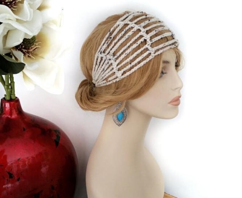 May include: A cream-colored, crocheted headband with a lattice design is worn on a mannequin with blonde hair. The mannequin also wears silver earrings with a turquoise stone. A red vase with white flowers is in the background.