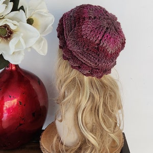 May include: A pink and purple knitted beanie hat with a textured design. The hat is on a mannequin head with long blonde hair.