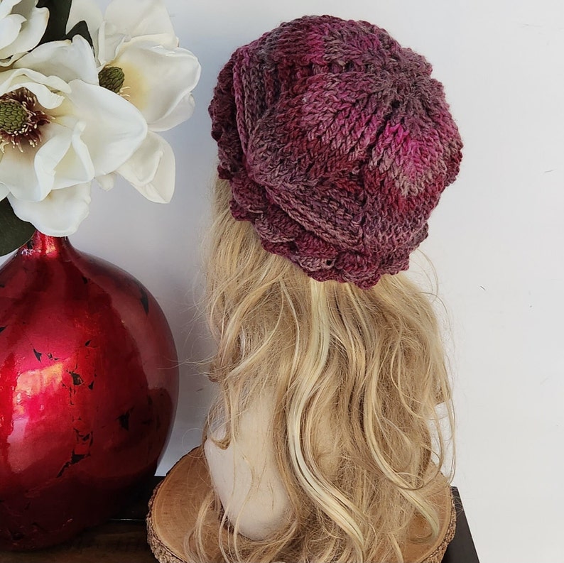May include: A pink and purple knitted beanie hat with a textured design. The hat is on a mannequin head with long blonde hair.