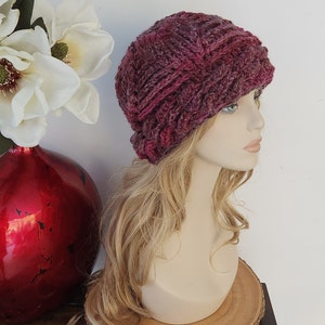 May include: A close-up of a pink and purple knitted beanie with a cable knit pattern. The beanie is on a mannequin with long blonde hair.