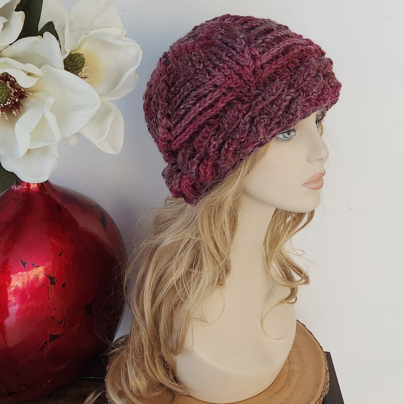 May include: A close-up of a pink and purple knitted beanie with a cable knit pattern. The beanie is on a mannequin with long blonde hair.