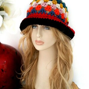 May include: A colorful crochet hat with a black brim and a red band. The hat features a patchwork design with various colors, including blue, yellow, pink, and green. The hat is displayed on a mannequin head with long blonde hair.