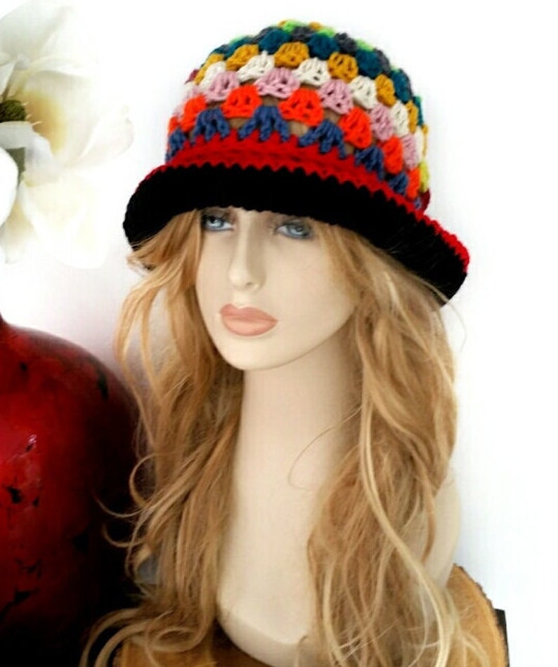 May include: A colorful crochet hat with a black brim and a red band. The hat features a patchwork design with various colors, including blue, yellow, pink, and green. The hat is displayed on a mannequin head with long blonde hair.