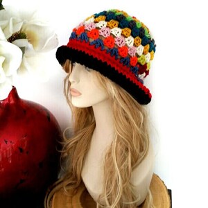 May include: A colorful crochet hat with a black brim and a red band. The hat features a patchwork design with various colors, including blue, orange, yellow, and green. The hat is displayed on a mannequin head with long, wavy blonde hair.