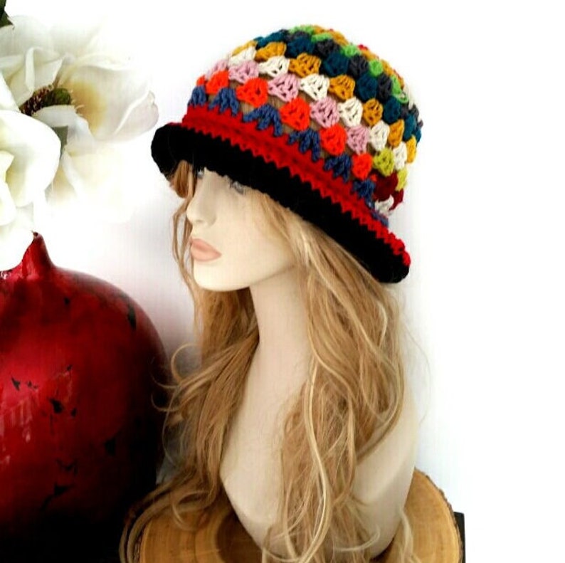 May include: A colorful crochet hat with a black brim and a red band. The hat features a patchwork design with various colors, including blue, orange, yellow, and green. The hat is displayed on a mannequin head with long, wavy blonde hair.