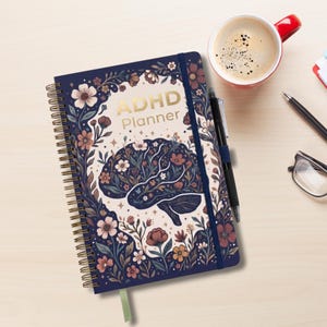 May include: A spiral-bound ADHD Planner with a dark blue cover featuring a central brain illustration filled with floral patterns in shades of pink, orange, and white. The text ADHD Planner is printed in gold. A black pen is attached to the side.