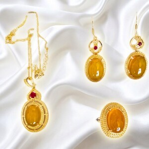 May include: A gold jewelry set consisting of a necklace, a pair of drop earrings, and a ring. Each piece features a large, oval, amber-colored cabochon stone surrounded by a textured gold frame, accented with a small, round, deep red gemstone.