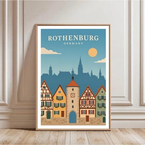 May include: A framed art print featuring a stylised illustration of Rothenburg, Germany. The image displays a row of colourful half-timbered houses with a central clock tower under a blue sky with a yellow sun and the text ROTHENBURG GERMANY.
