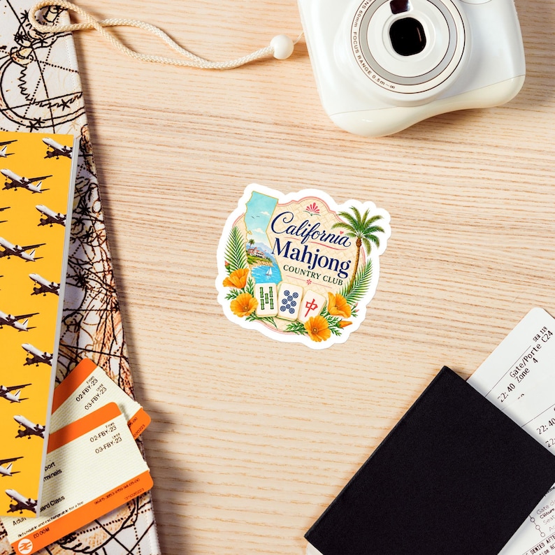 May include: A die-cut sticker featuring the text California Mahjong Country Club, a map of California, a palm tree, orange poppies, and three mahjong tiles. The design is framed by tropical foliage and a coastal scene with a blue ocean and sky.