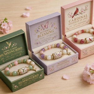 May include: Three beaded bracelets in individual boxes. One green box labeled Spring Garden Hummingbird, one purple box labeled Butterfly Blessing, and one pink box labeled Cardinal Messenger of Love. Each bracelet features a central ceramic bead.