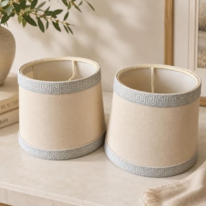 May include: Two cylindrical lamp shades in a neutral beige fabric, each featuring a light blue trim at the top and bottom edges. The trim is decorated with a repeating geometric Greek key pattern. The interior of each shade is a plain off-white color.