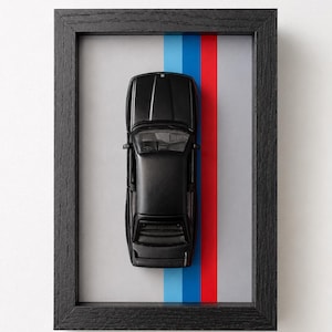 Pode incluir: Um carro em miniatura preto montado dentro de uma moldura de madeira preta. O fundo apresenta uma superfície cinza-claro com listras verticais azul-claro, azul-escuro e vermelhas posicionadas atrás do carro, visto de cima.