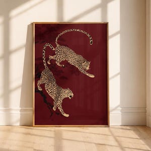 May include: A rectangular framed wall art print featuring two leopards with black spots on a deep burgundy background. The leopards are depicted in a dynamic pose, one leaping above the other, with dark tree branches visible in the background.