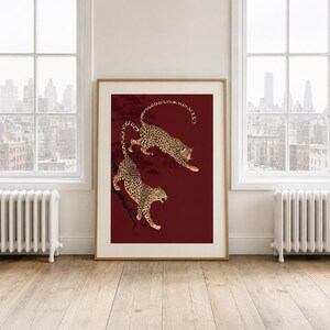 May include: A framed vertical wall art print featuring two spotted leopards in a dynamic pose against a deep burgundy background. The leopards are depicted with black spots on a tan coat, positioned as if climbing or leaping on dark branches.