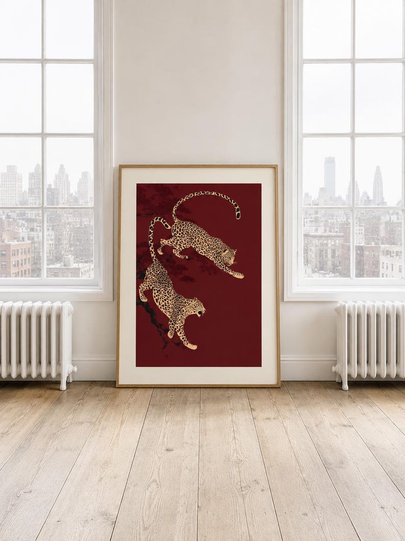 May include: A framed vertical wall art print featuring two spotted leopards in a dynamic pose against a deep burgundy background. The leopards are depicted with black spots on a tan coat, positioned as if climbing or leaping on dark branches.