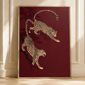 May include: A framed rectangular wall art print featuring two spotted leopards in a dynamic pose against a deep burgundy background. The leopards are depicted with black spots on a cream-colored coat, positioned as if leaping or climbing.
