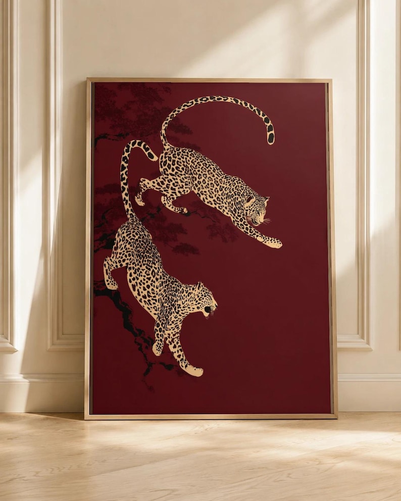 May include: A framed rectangular wall art print featuring two spotted leopards in a dynamic pose against a deep burgundy background. The leopards are depicted with black spots on a cream-colored coat, positioned as if leaping or climbing.