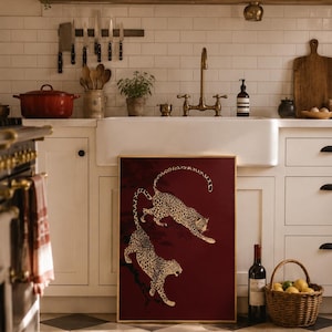 May include: A rectangular framed wall art piece featuring two leopards in a dynamic leaping pose against a deep burgundy background. The leopards are depicted with detailed black spots on a tan coat, positioned as if moving through dark tree branches.