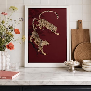 May include: A rectangular wall art print in a white frame featuring two leopards with black spots on a deep burgundy background. The leopards are depicted in motion, positioned as if climbing or leaping among dark, silhouetted tree branches.