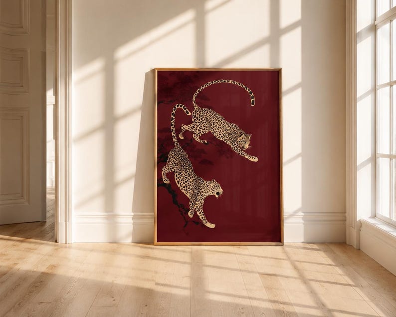 May include: A rectangular framed wall art print featuring two leopards with black spots on a deep burgundy background. The leopards are depicted in a dynamic pose, one leaping above the other, with dark tree branches visible in the background.