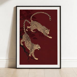 May include: A framed wall art print featuring two leopards with tan fur and black spots against a deep burgundy background. The leopards are depicted in dynamic poses, one leaping and one descending, with dark silhouettes of tree branches visible.