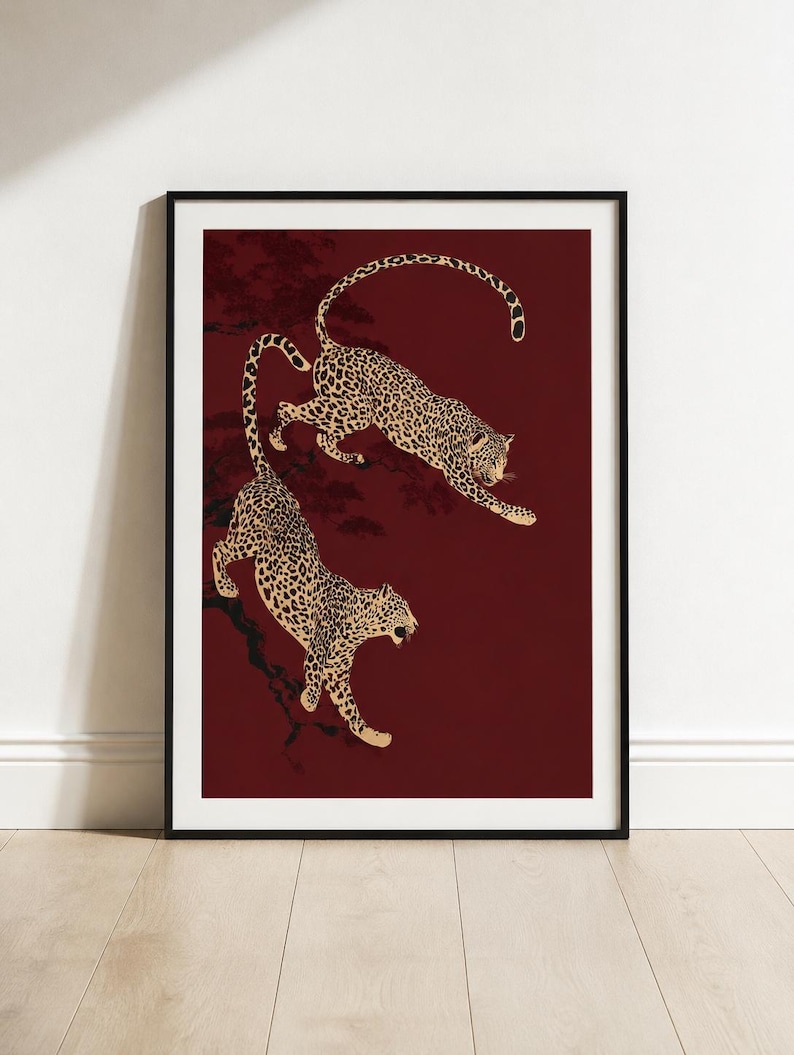 May include: A framed wall art print featuring two leopards with tan fur and black spots against a deep burgundy background. The leopards are depicted in dynamic poses, one leaping and one descending, with dark silhouettes of tree branches visible.