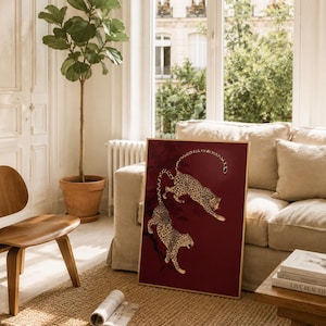 May include: A rectangular framed wall art piece featuring two gold-colored leopards depicted in a dynamic, leaping pose against a deep, solid burgundy red background. The leopards are illustrated with detailed black spots and are positioned diagonally.