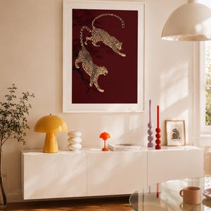May include: A framed vertical wall art print featuring two leopards with spotted fur climbing on dark branches against a deep burgundy background. The artwork is displayed in a white frame, showcasing a detailed illustration of the wild cats.