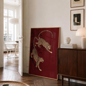 May include: A large rectangular framed wall art piece featuring a deep red background with two illustrated leopards climbing on dark tree branches. The leopards are depicted with gold and black spotted fur, positioned one above the other.