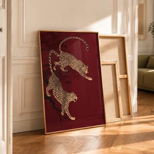 May include: A rectangular framed wall art piece featuring two leopards with gold and black spotted fur against a deep burgundy red background. The leopards are depicted in dynamic poses, one leaping above the other, set against dark tree branches.