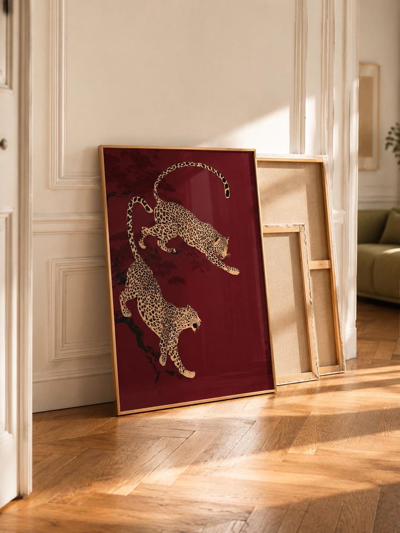 May include: A rectangular framed wall art piece featuring two leopards with gold and black spotted fur against a deep burgundy red background. The leopards are depicted in dynamic poses, one leaping above the other, set against dark tree branches.