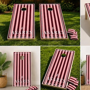 Könnte beinhalten: Ein Cornhole-Spielset bestehend aus zwei rechteckigen Holzbrettern mit vertikalem Streifenmuster in Rosa- und Dunkelrottönen. Jedes Brett hat ein kreisförmiges Loch oben und den weißen Schriftzug "The ASARES EST 2016" auf der Vorderseite.