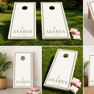 Op de afbeelding: Een set van twee witte cornhole-borden met donkergroene randlijnen en de tekst "The ASARES EST. 2024" in het donkergroen gedrukt. De borden hebben een cirkelvormig gat aan de bovenkant en worden vergezeld door kleine lichtroze en witte zitzakjes.