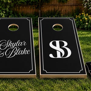 Könnte beinhalten: Zwei schwarze Cornhole-Boards mit weißen Rändern und kreisförmigen Löchern. Ein Board zeigt den Text Skylar & Blake in weißer Schreibschrift, während das andere ein großes weißes SB-Monogramm aufweist. Vier passende schwarze Sitzsäcke mit SB-Logo sind enthalten.