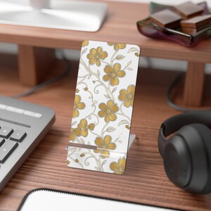 May include: A rectangular phone stand featuring a white background with a repeating pattern of gold-colored flowers and thin, swirling vines. The stand has a clear acrylic base and a horizontal support ledge, displaying a floral design throughout.