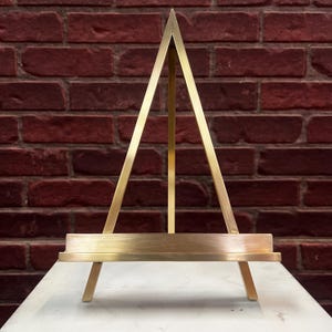 May include: A triangular gold-toned metal display easel featuring a horizontal ledge for supporting items. The structure consists of two angled side supports meeting at a top point, a central vertical brace, and a flat base shelf with a smooth finish.