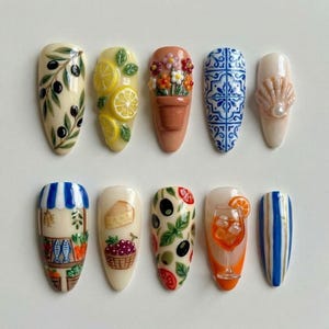 May include: A set of ten almond-shaped press-on nails featuring detailed 3D and painted designs including olive branches, lemon slices, a potted flower, blue and white tiles, a seashell, a market stall, cheese, grapes, salad, a cocktail, and stripes.