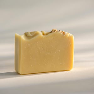 May include: A rectangular bar of pale yellow soap with a smooth, solid texture. The top edge features a slightly uneven, molded surface with a small cluster of light brown, textured botanical inclusions embedded on the upper right corner.