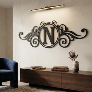 May include: A black metal wall art piece featuring a central circular frame enclosing the capital letter N, flanked by symmetrical scrolling filigree designs. The decorative sign is mounted on a wall beneath a horizontal linear light fixture.