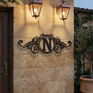 May include: A black metal wall sign featuring a central circular frame with the letter N inside, flanked by symmetrical scrollwork designs. The sign is mounted on a light brown stone wall between two lit copper-toned outdoor wall lanterns.