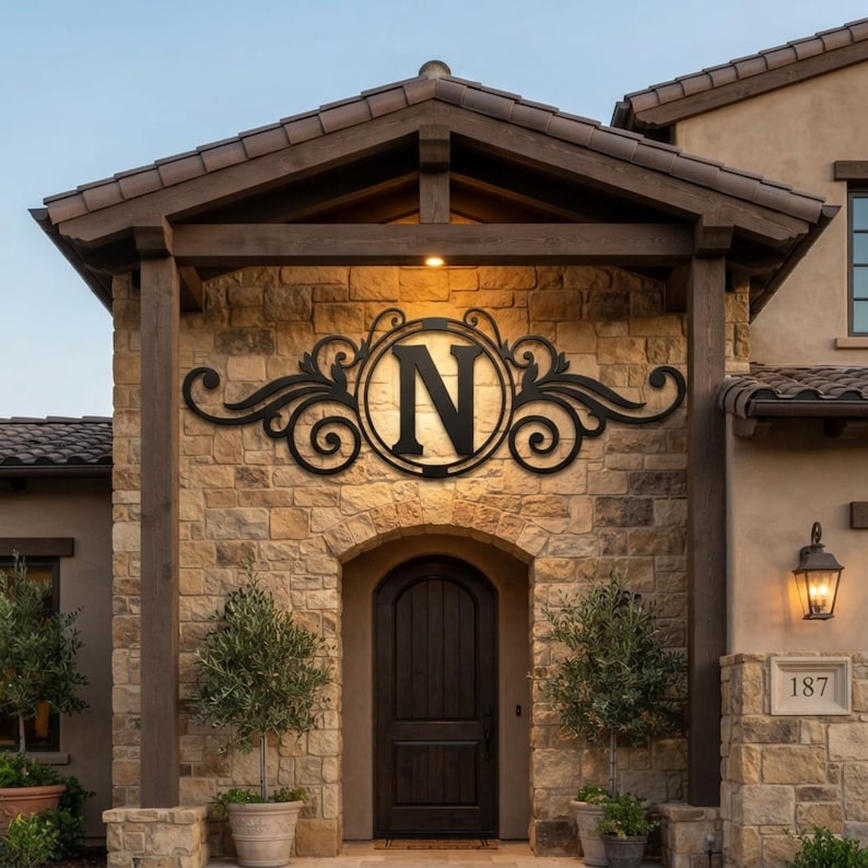 May include: A black metal wall sign featuring a large capital letter N centred within a circular frame, surrounded by decorative scrollwork and flourishes. The sign is mounted on a light brown stone exterior wall above a dark wooden front door.